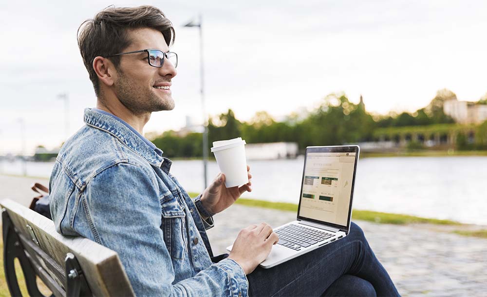 Man seating un a bench while drinking coffee and watching a computer.